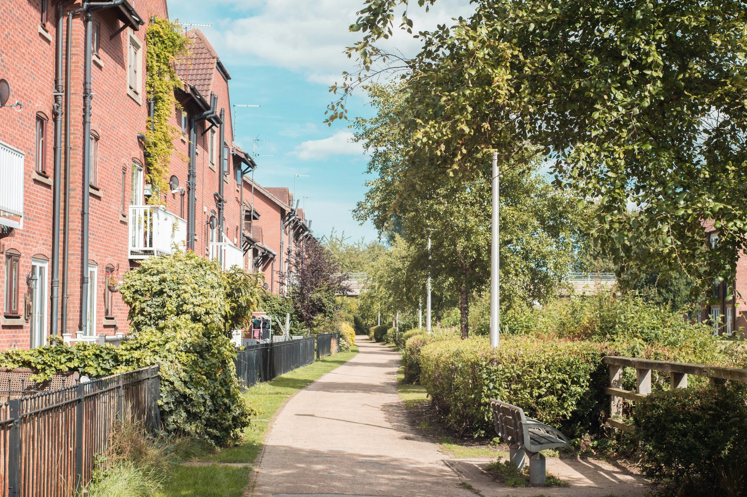 A tranquil pathway lined with trees and bushes, bordered by red brick residential buildings on one side. The path features a bench and is bathed in sunlight, with a clear blue sky overhead.