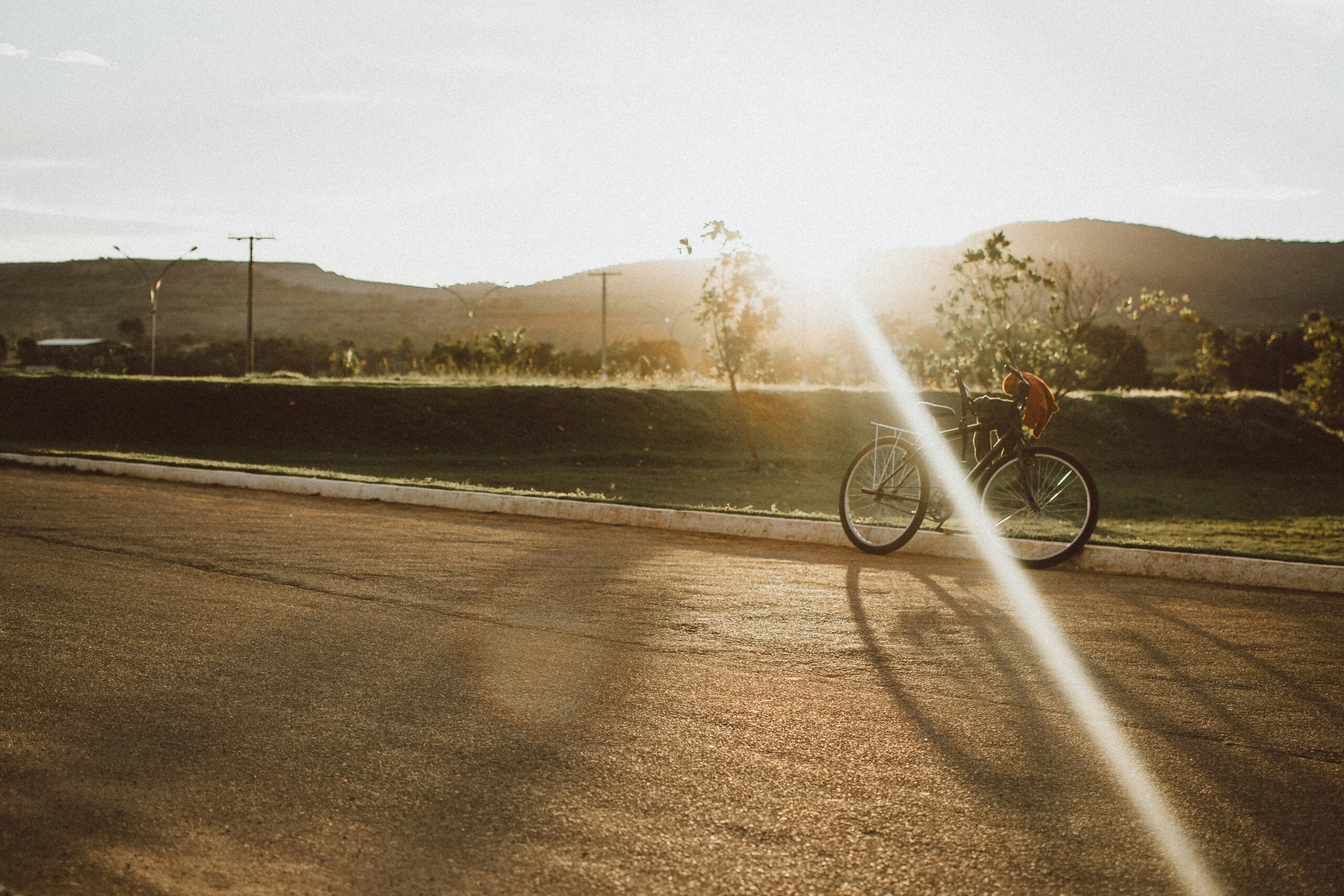 A bicycle resting on a quiet road at sunset, with hills and trees in the background, casting long shadows. 